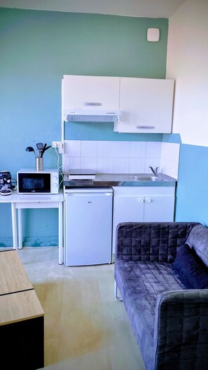 Cozinha compacta com micro-ondas, torradeira e mini geladeira. Na sala, há um sofá e uma mesa.
