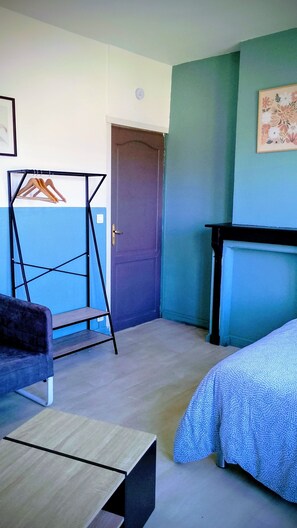 Um quarto com uma cama, uma cadeira, uma mesa pequena, uma porta e um quadro na parede.