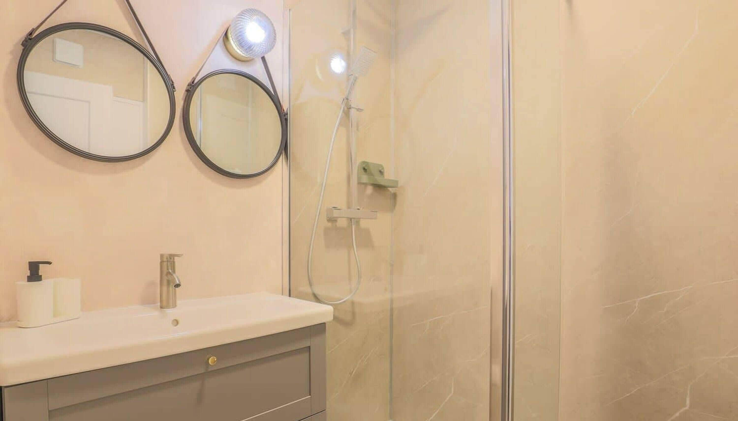 A bathroom with a glass shower enclosure, two round mirrors, a white sink with a faucet, and a soap dispenser.
