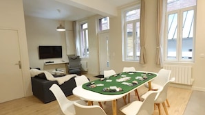 Una habitación con una mesa de póker verde, sillas blancas, un sofá gris, un televisor de pantalla plana y ventanas grandes con cortinas.