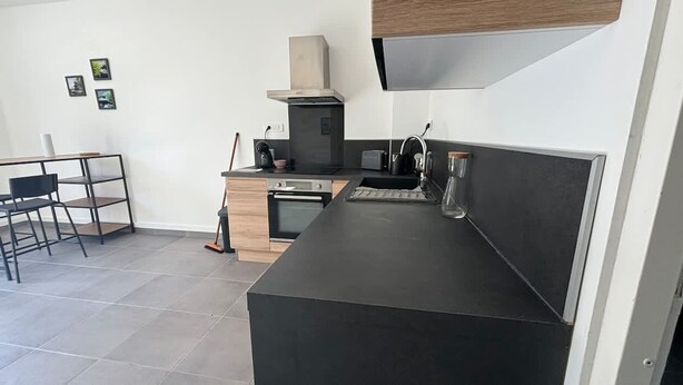 A modern kitchen with a black countertop, a built-in oven, a stove, and a sink. There is a dining area with a chair and a table. - Le Rietz 3 T2 Proche Gare et Parc (Arras)