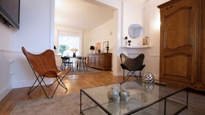 Ein Wohnzimmer mit einem Glastisch, zwei Ledersesseln, einer Holzkommode und einem Essbereich im Hintergrund.