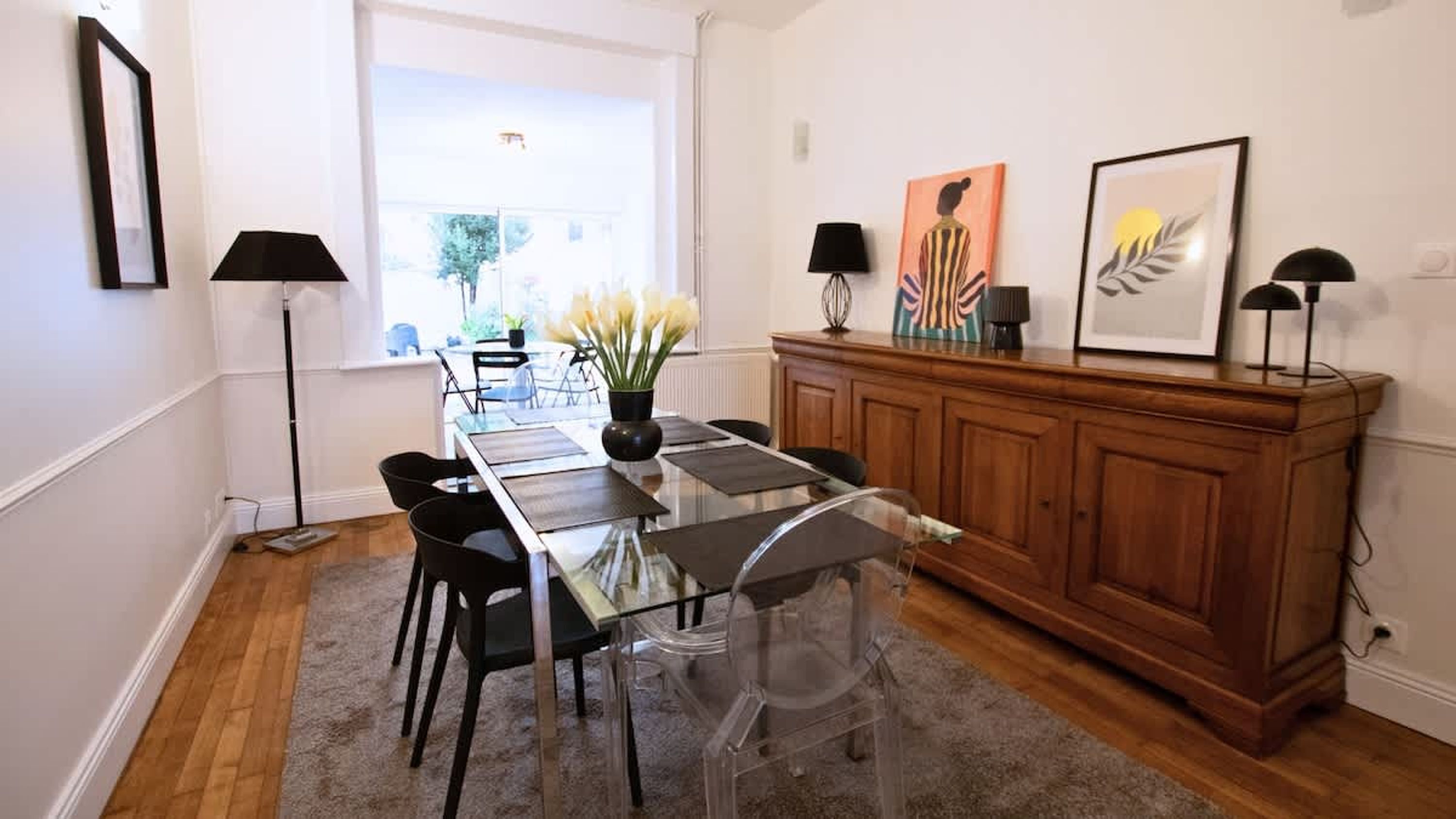 Een eetgedeelte met een glazen tafel, zwarte stoelen, een houten kast en kunstwerken aan de wanden.