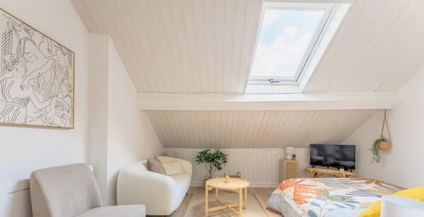 A room with a sloped ceiling, a skylight, a bed, a TV, a plant, and a framed picture on the wall. - Les Combles d Eugene Proche Bollaert 2pers (Lens)