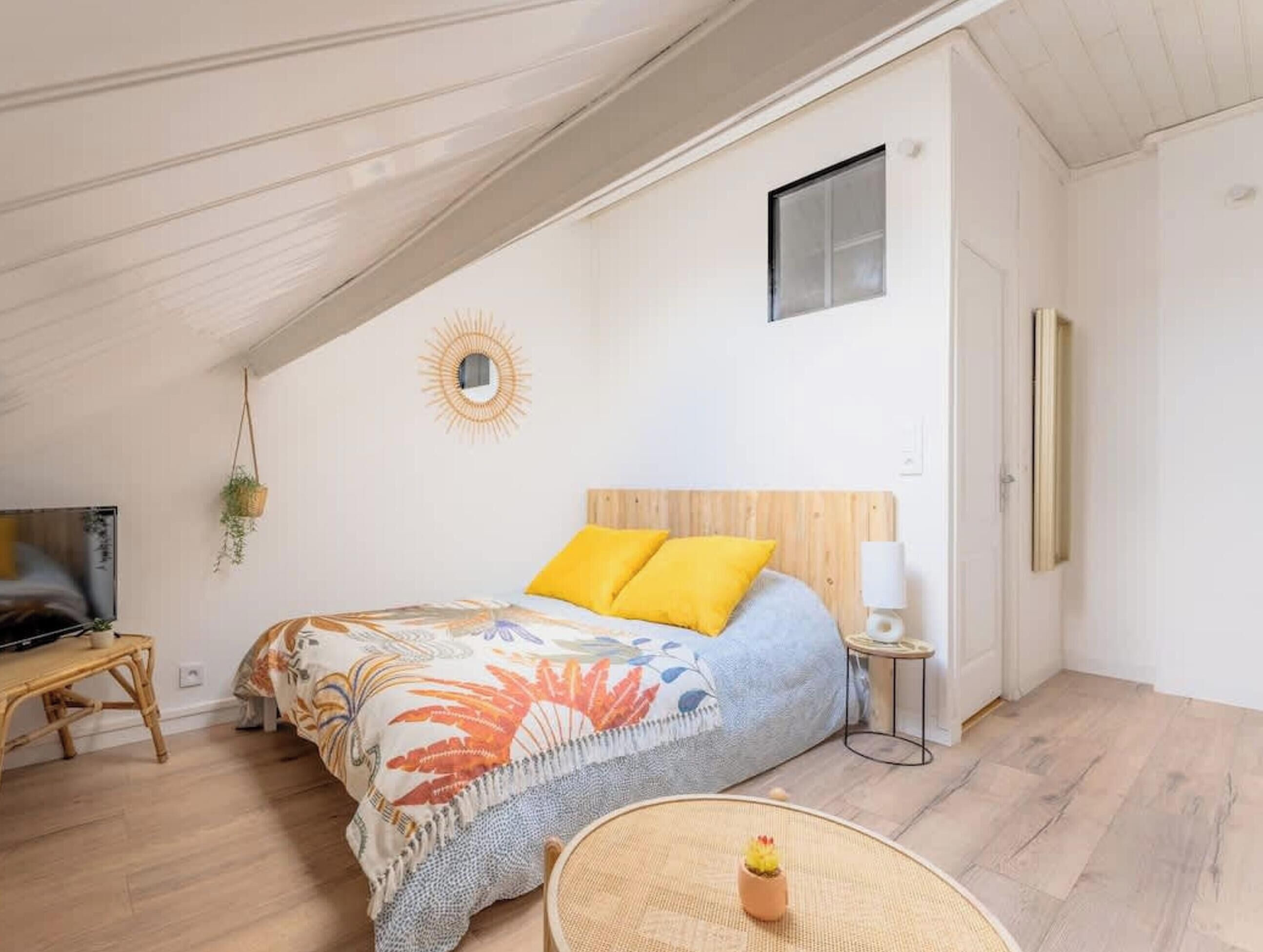 A bedroom with a sloped ceiling, a bed with a wooden headboard, a small window, a wall-mounted mirror, a bedside table with a lamp, and a hanging plant.