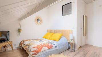 A bedroom with a sloped ceiling, a bed with a wooden headboard, a small window, a wall-mounted mirror, a bedside table with a lamp, and a hanging plant.