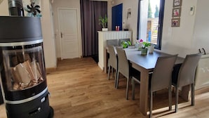 A cozy room with a wood-burning stove, a dining table with chairs, a small kitchen area, and a door leading to another room. - La maison aux palmiers, swimming pool, spa (Sorigny)