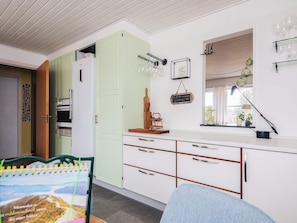 A kitchen with white cabinets, a green door, and a window with blinds. There are wine glasses on a rack and a chalkboard on the wall. - 4 Person Holiday Home in Norre Nebel (Norre Nebel)