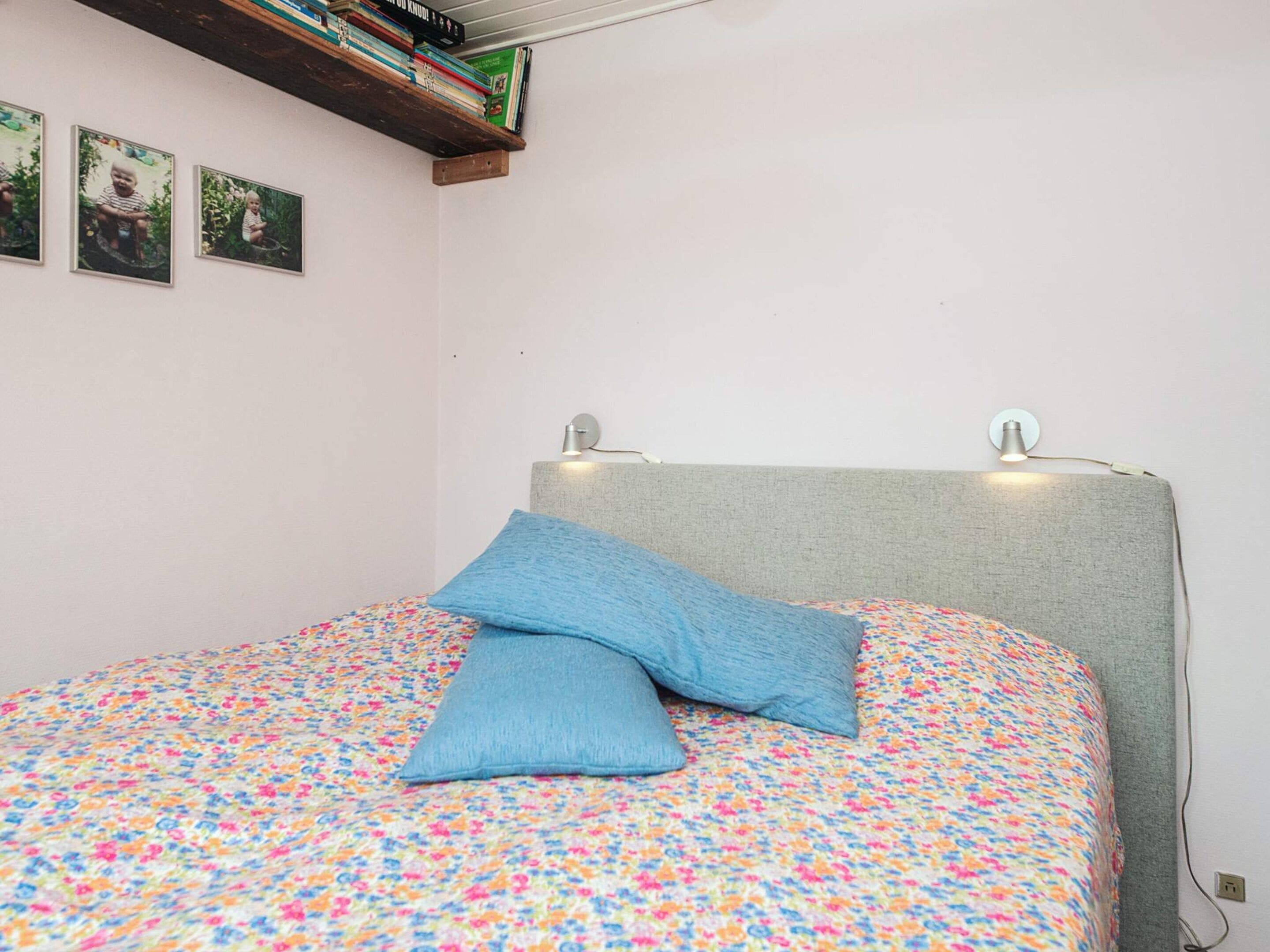 A bedroom with a bed, a pillow, a shelf with books, and a wall-mounted light.