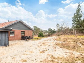 A single-story brick building with a sloped roof, a small attached garage, and a dirt driveway leading to a grassy area. - 4 Person Holiday Home in Norre Nebel (Norre Nebel)