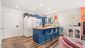 A modern kitchen with blue cabinets, a white refrigerator, a wooden island with bar stools, and a small seating area with a pink chair. - Daylight Cove Lakefront Chic (Lake Ozark)