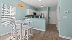 A modern kitchen with white cabinets, a stainless steel refrigerator, and a dining area with a white table and chairs. - Cross Gate 104 - End Unit, Lake View, Pool (Surfside)