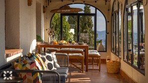 Un patio soleggiato con un tavolo apparecchiato per la colazione, un divano con cuscini colorati e vista sul mare attraverso ampie finestre.
