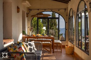 A sunlit patio with a table set for breakfast, a sofa with colorful cushions, and a view of the sea through large windows. - Villa ROC by Abahana Villas (Benissa)