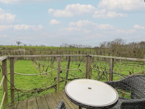 Eine Holzdielen-Terrasse mit rundem Tisch und Korbstühlen, mit Blick auf einen Weinberg.