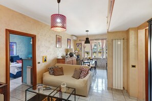 Een woonkamer met een bank, een glazen salontafel, een rode hanglamp, een keuken met een eettafel, en een gang die leidt naar een slaapkamer.