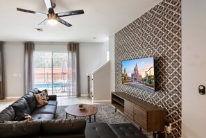 A modern living room with a flat-screen TV mounted on a patterned wall, a ceiling fan, a sofa with decorative pillows, a coffee table, and a staircase leading to an upper level. - Sweet Retreat At Bella Vida (Kissimmee)