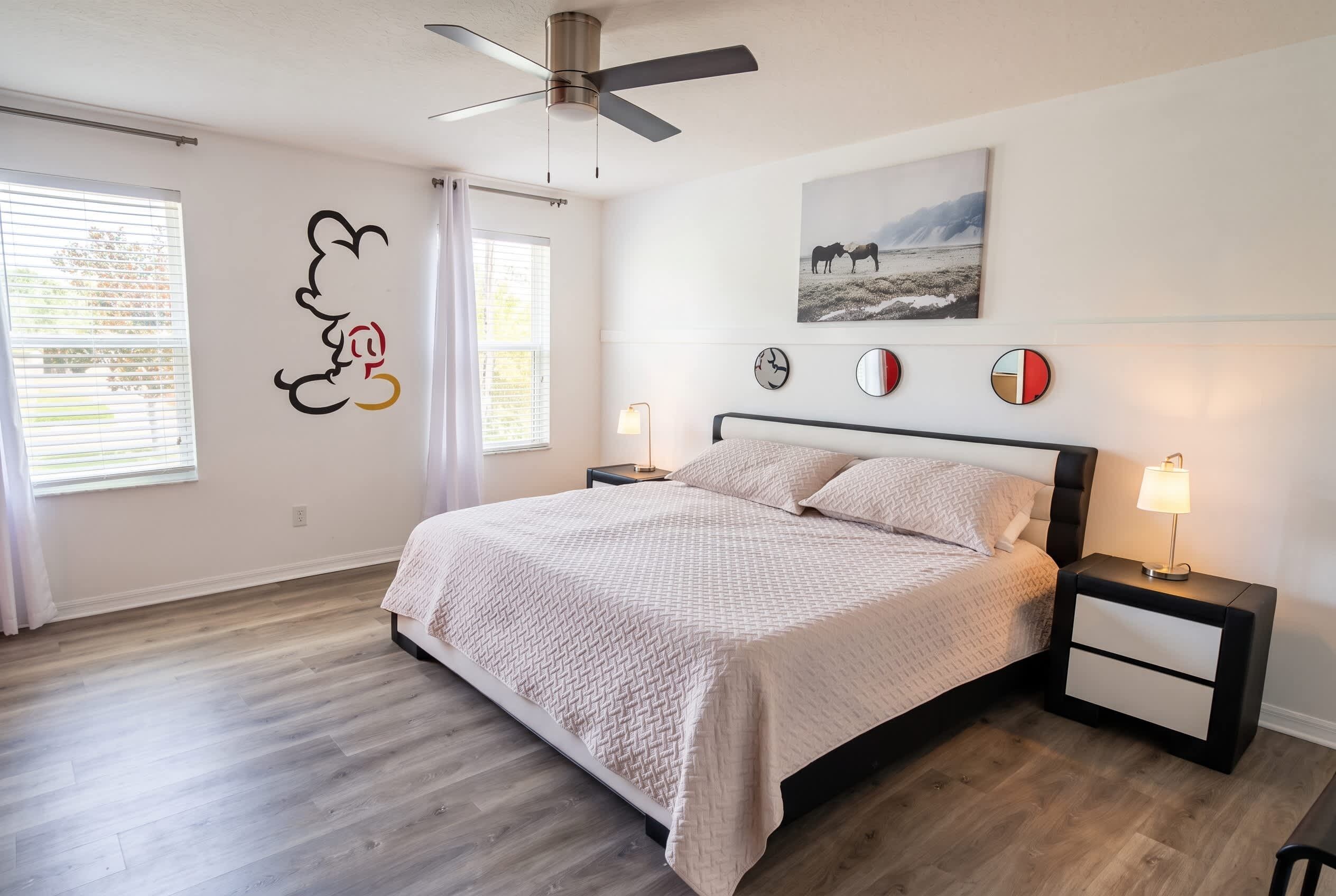 Quarto com cama, mesas de cabeceira, ventoinha no teto e obra de arte na parede.