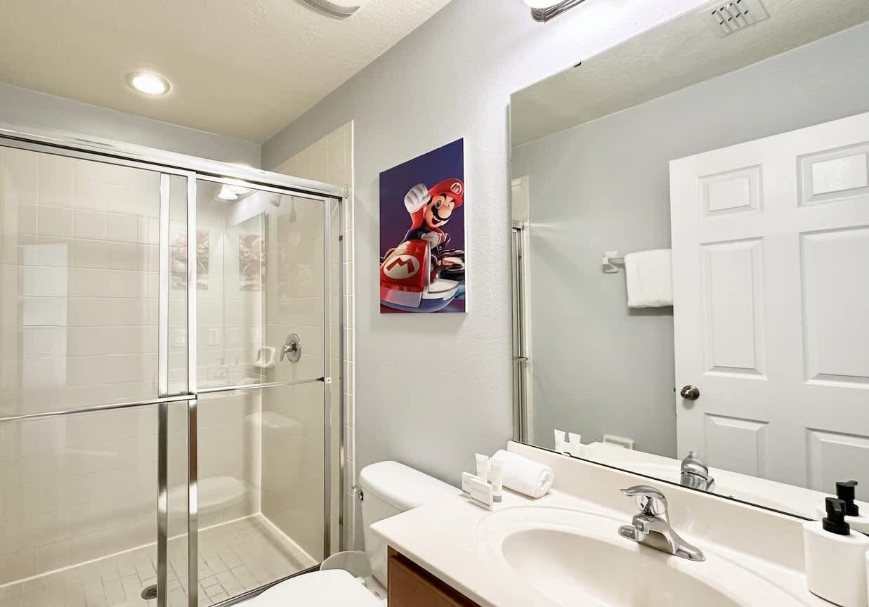 A modern bathroom with a glass shower enclosure, a white sink with a mirror, and a poster of a video game character.