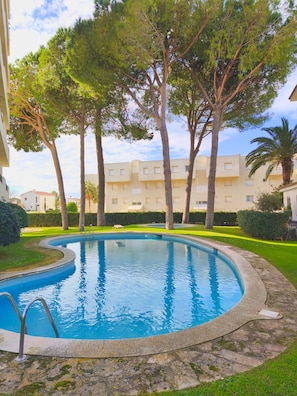 A swimming pool with a stone border, surrounded by trees and a building with balconies. - Iona (L'Escala)