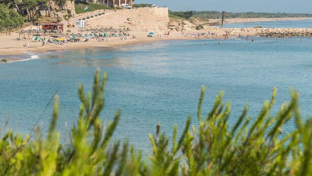 A beach with clear blue water, a sandy shore, and a stone wall in the background. - Iona (L'Escala)