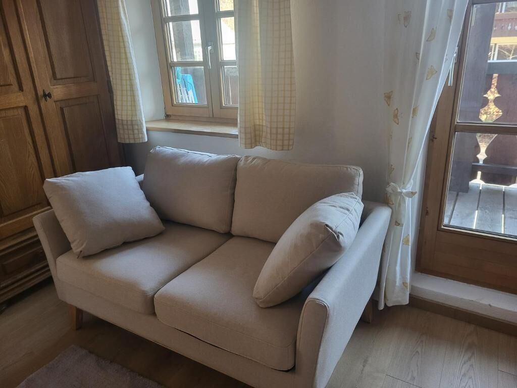 A beige sofa with cushions, a wooden wardrobe, a window with curtains, and a view of a balcony.