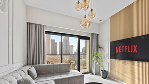 A modern living room with a grey sofa, a large window with curtains, a potted plant, and a wall-mounted TV displaying the Netflix logo. - Luxury Burj Royale 2BR Burj Khalifa & Fountain View With Balcony by Evoke (Dubai)