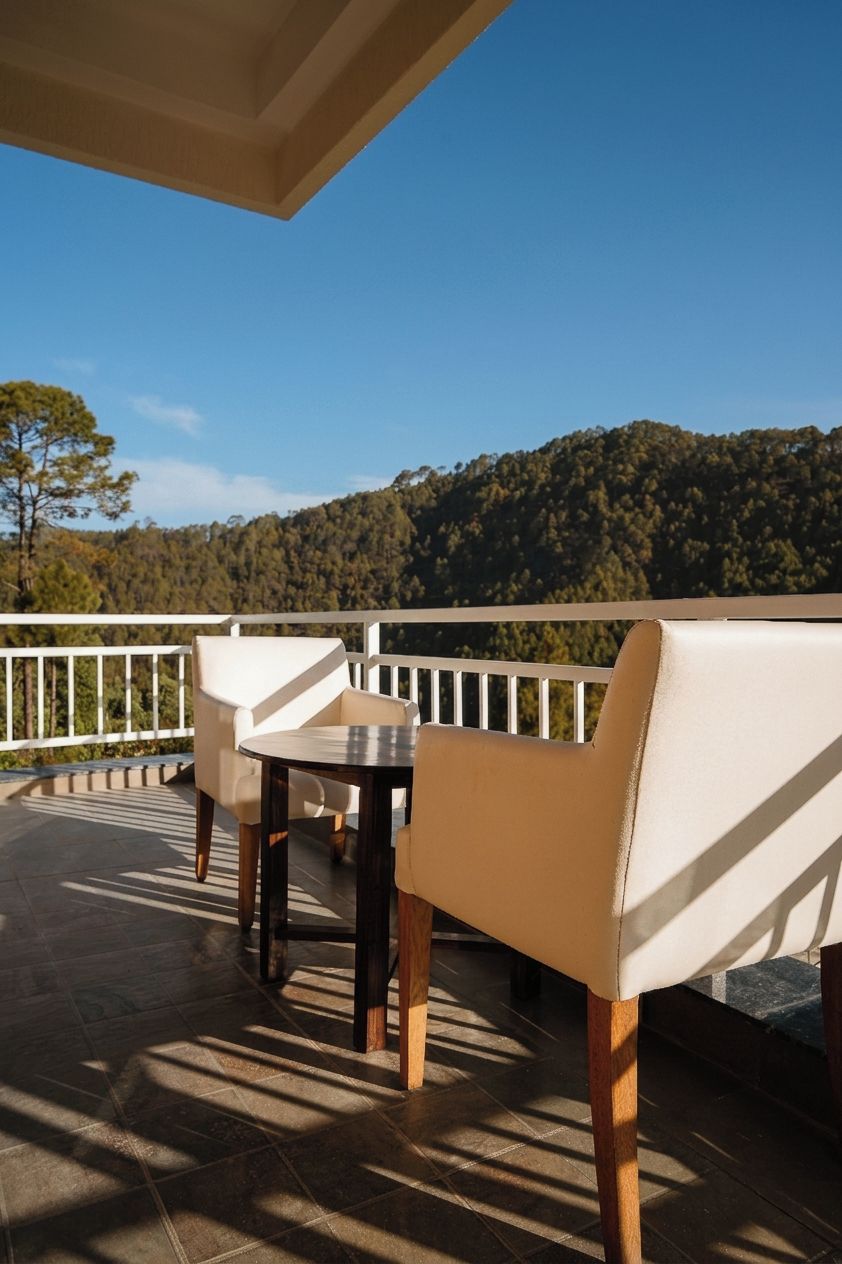 A terrace with a table and chairs, a white railing, and a forested hill in the background.