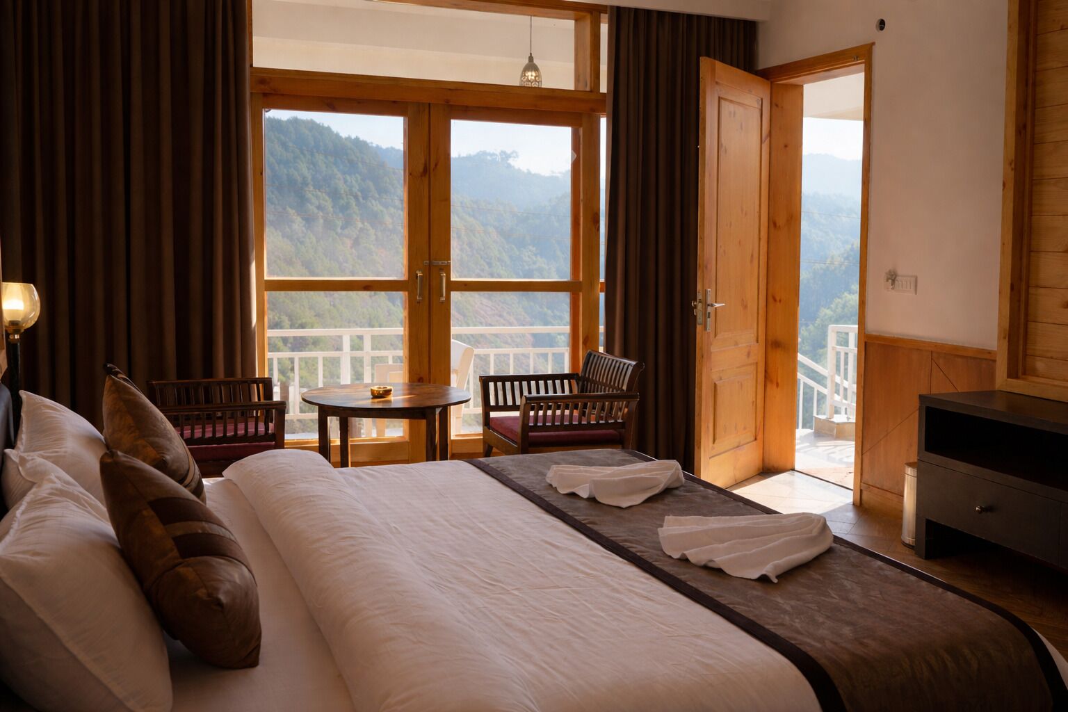 A hotel room with a bed, a table, two chairs, a desk, and a balcony with a mountain view.