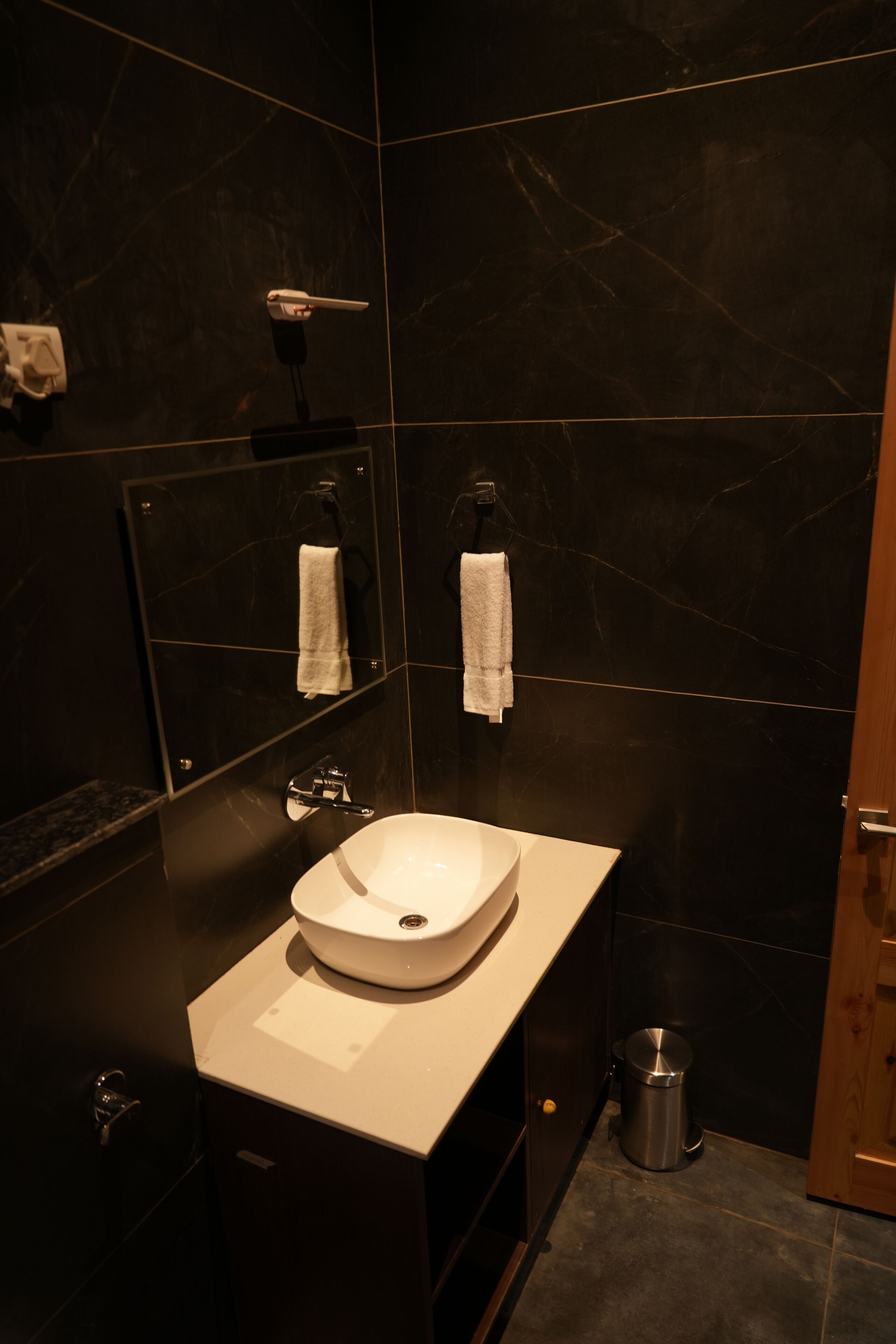 Uma casa de banho moderna com uma pia branca, paredes de mármore escuro, um espelho e uma pequena lixeira.