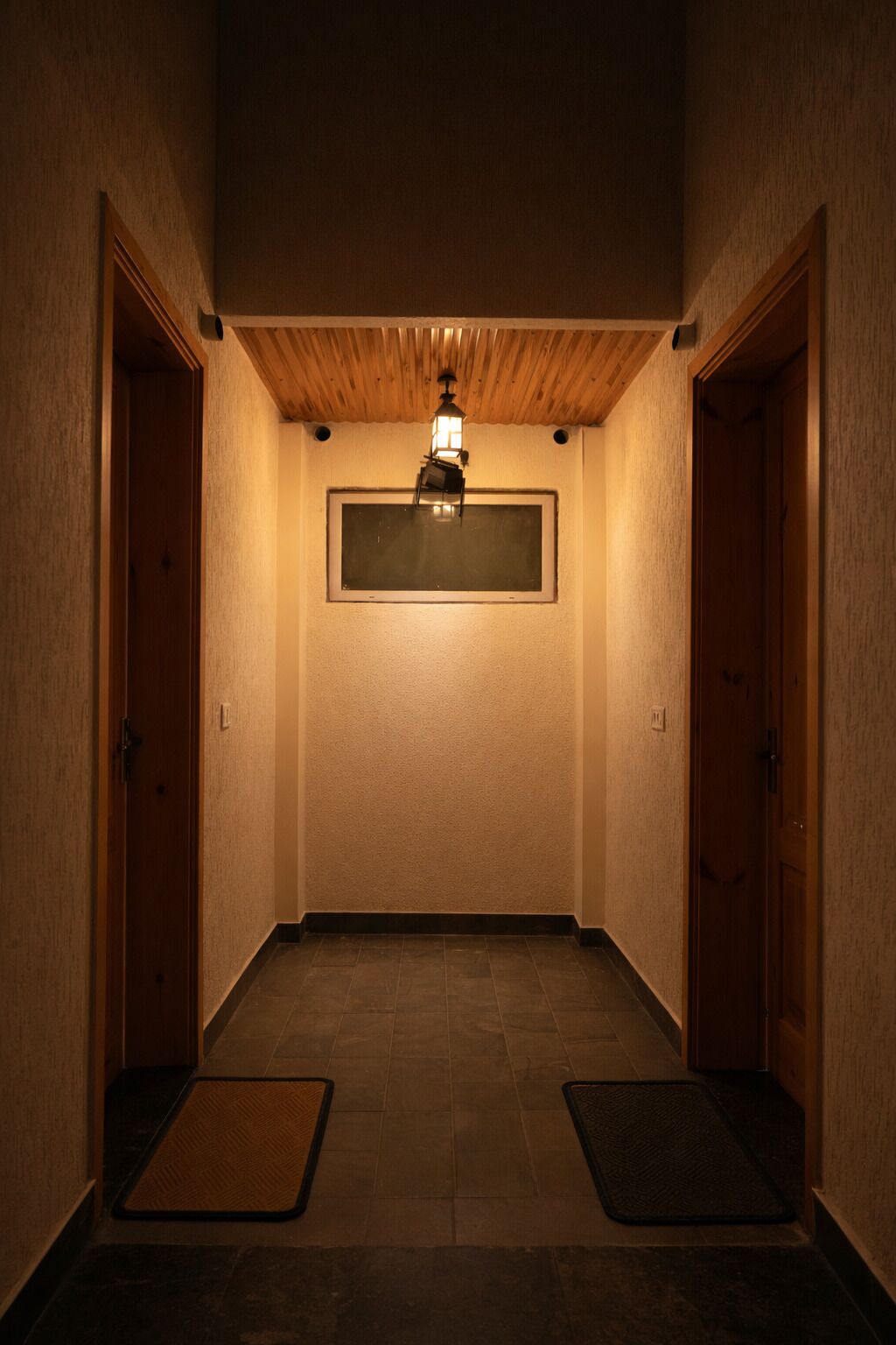 Un pasillo con puertas de madera, un muro con textura, un ventanal rectangular y una lámpara de pie.