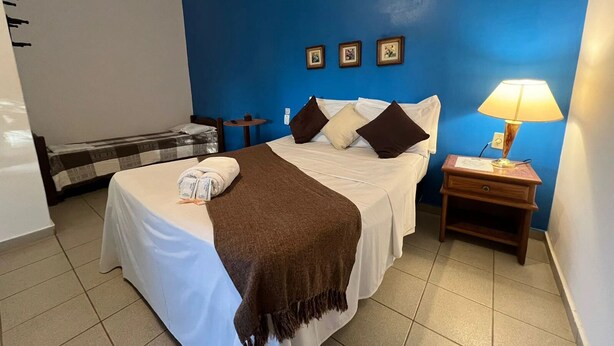 Ein Hotelzimmer mit Bett, Nachttisch mit Lampe, einem kleinen Tisch mit Stuhl und einer blauen Wand mit gerahmten Bildern.