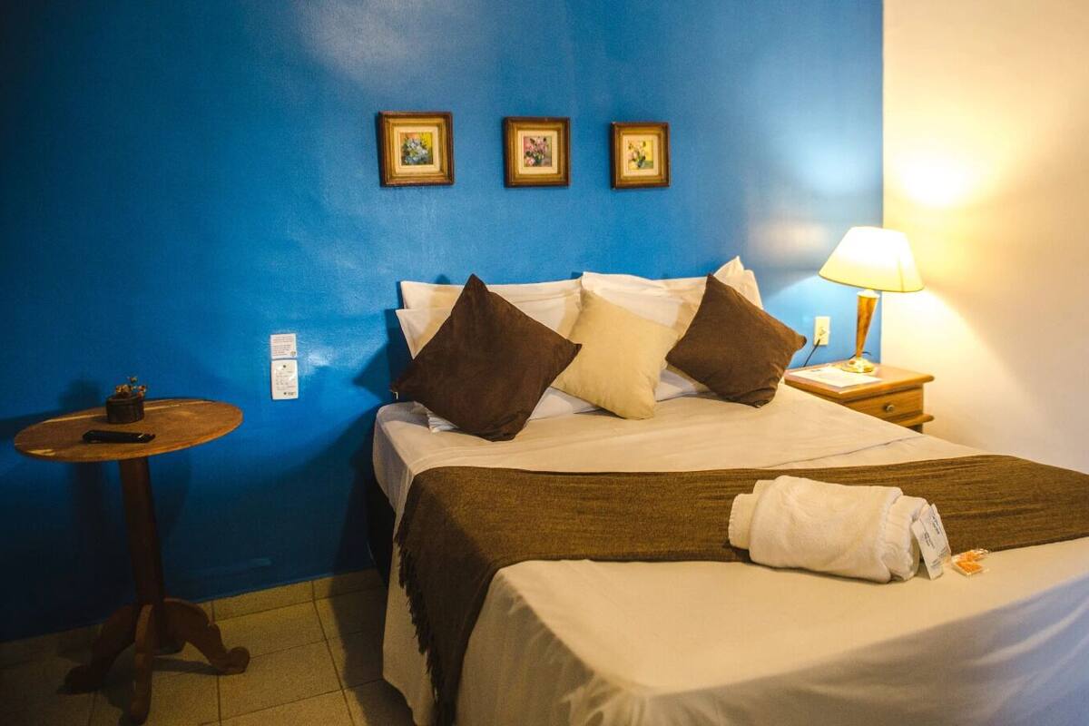 Quarto de hotel com uma cama, mesa de cabeceira, abajur e três quadros na parede.