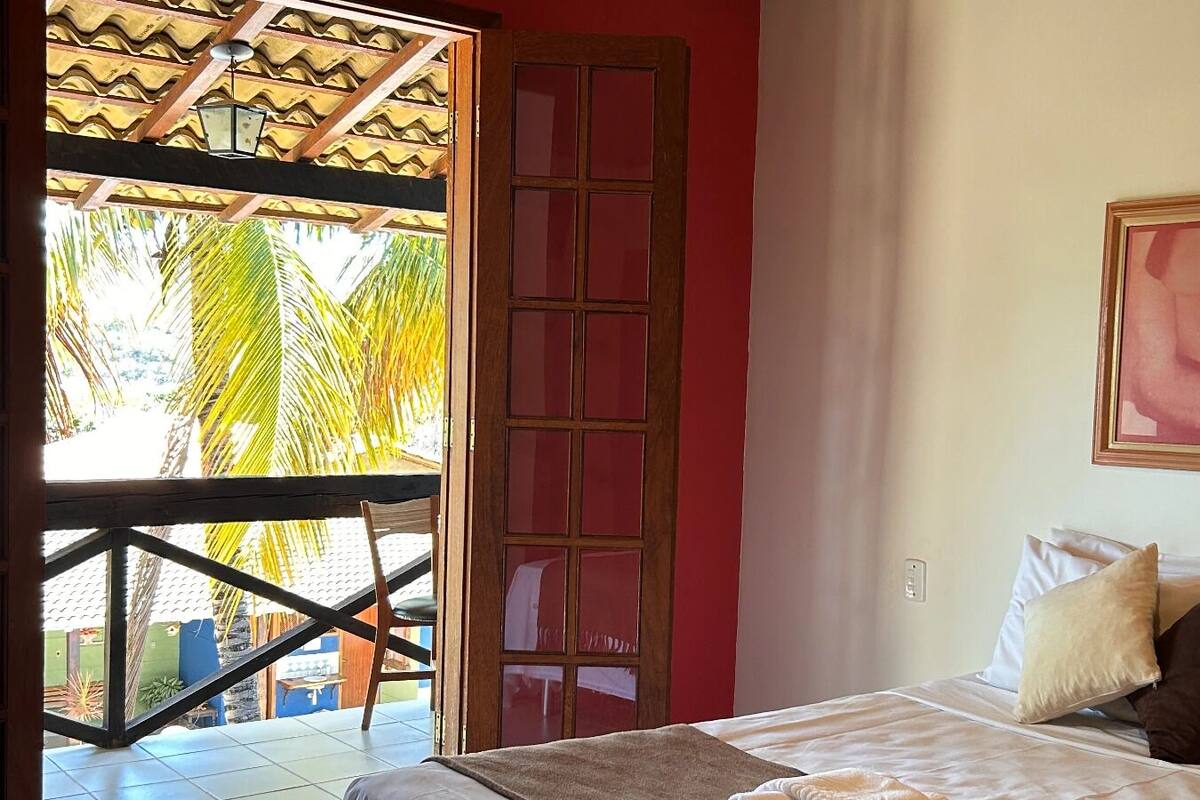 Quarto com uma cama, janela com vista para palmeiras, porta de madeira e um quadro pendurado na parede.