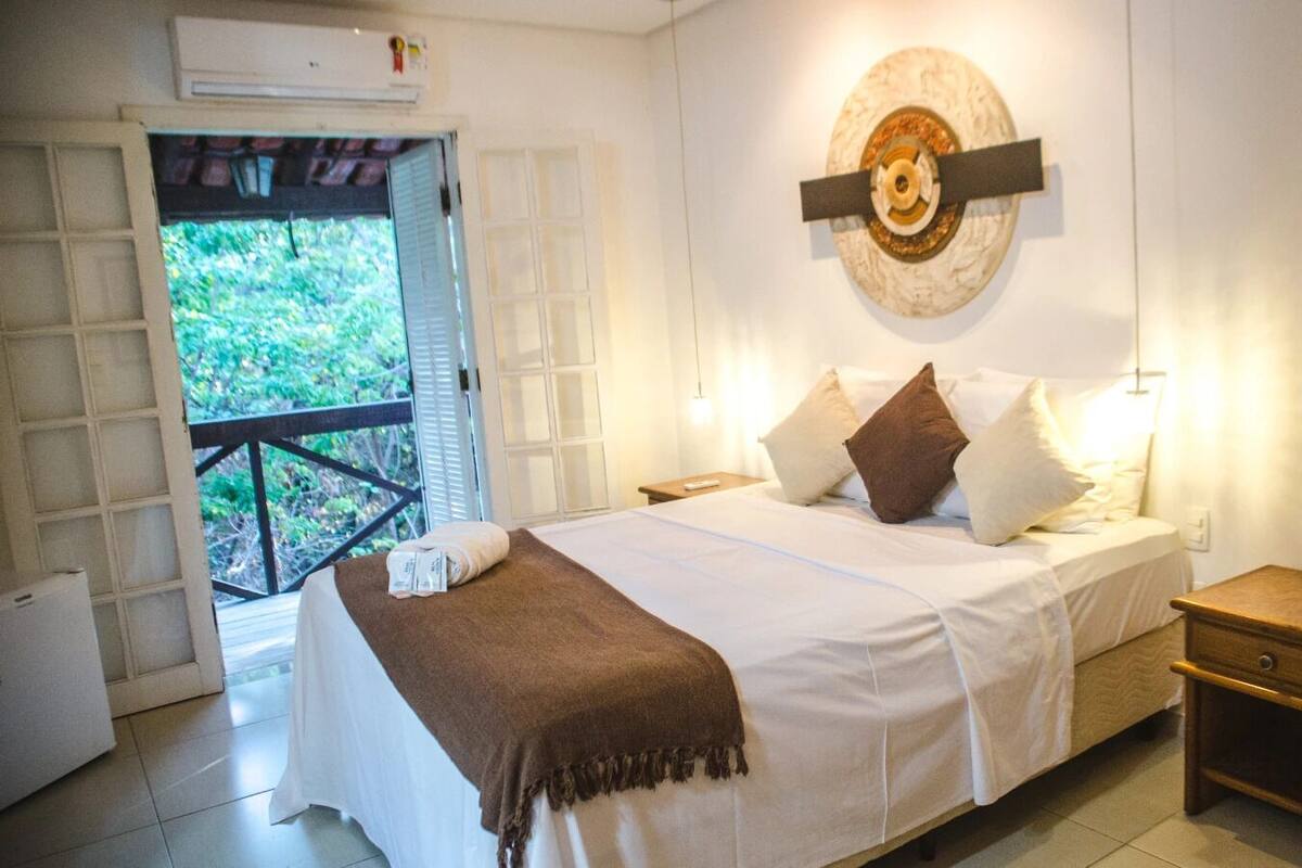 Quarto com cama, mesa de cabeceira e vista para o verde através de uma porta aberta.