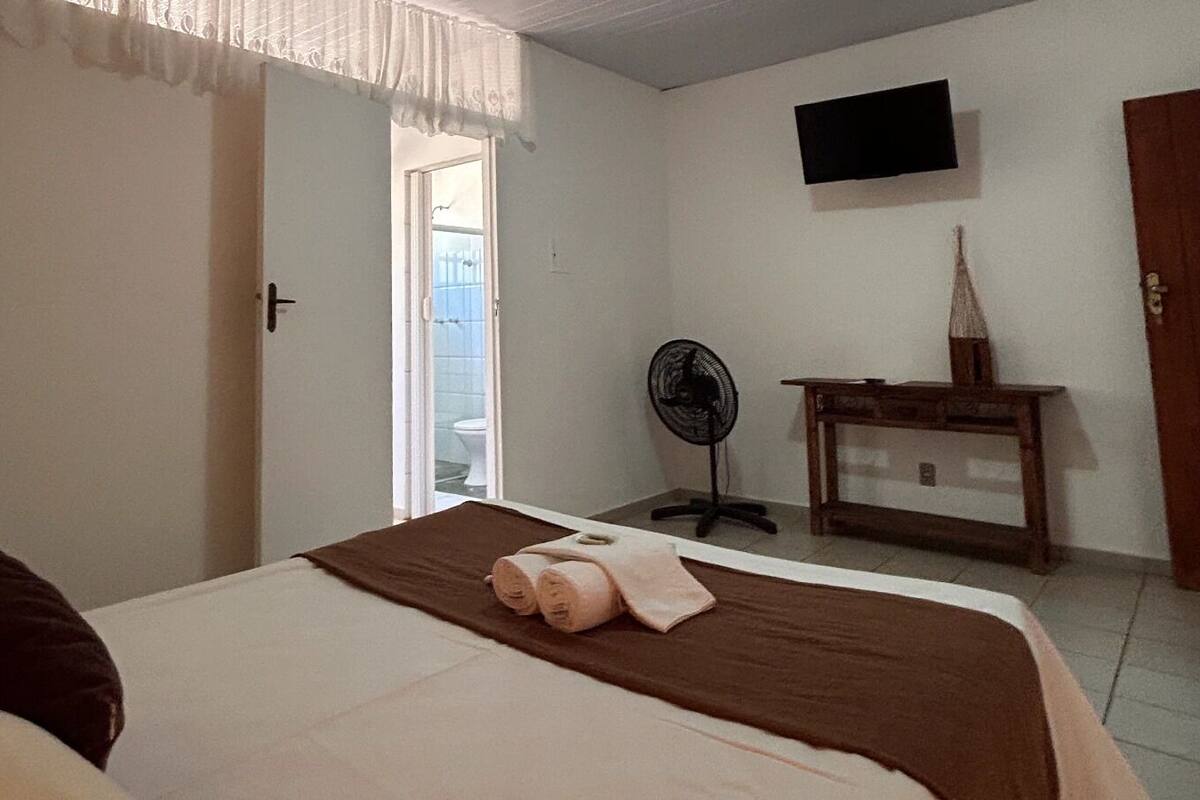Um quarto com uma cama, mesa de cabeceira, ventilador, televisão e uma porta para uma varanda.