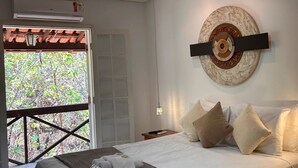 Una camera da letto con un letto, cuscini, una sveglia appesa al muro e vista su alberi attraverso una porta.
