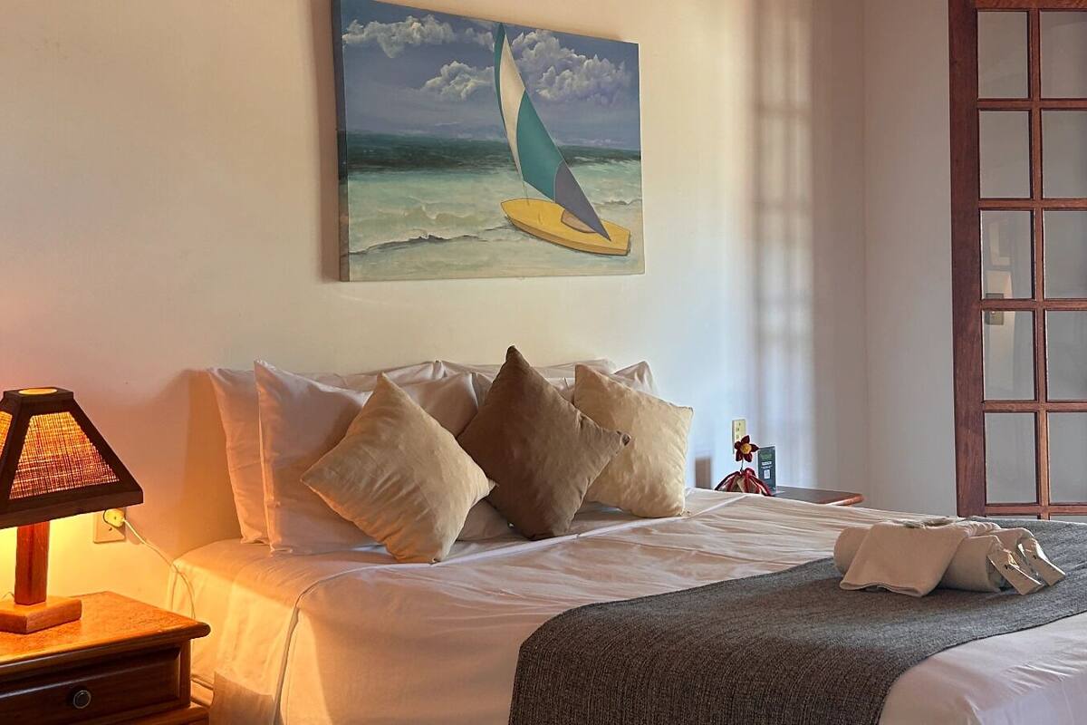 Quarto de hotel com cama, travesseiros, luminária, quadro de um veleiro e vista para palmeiras.