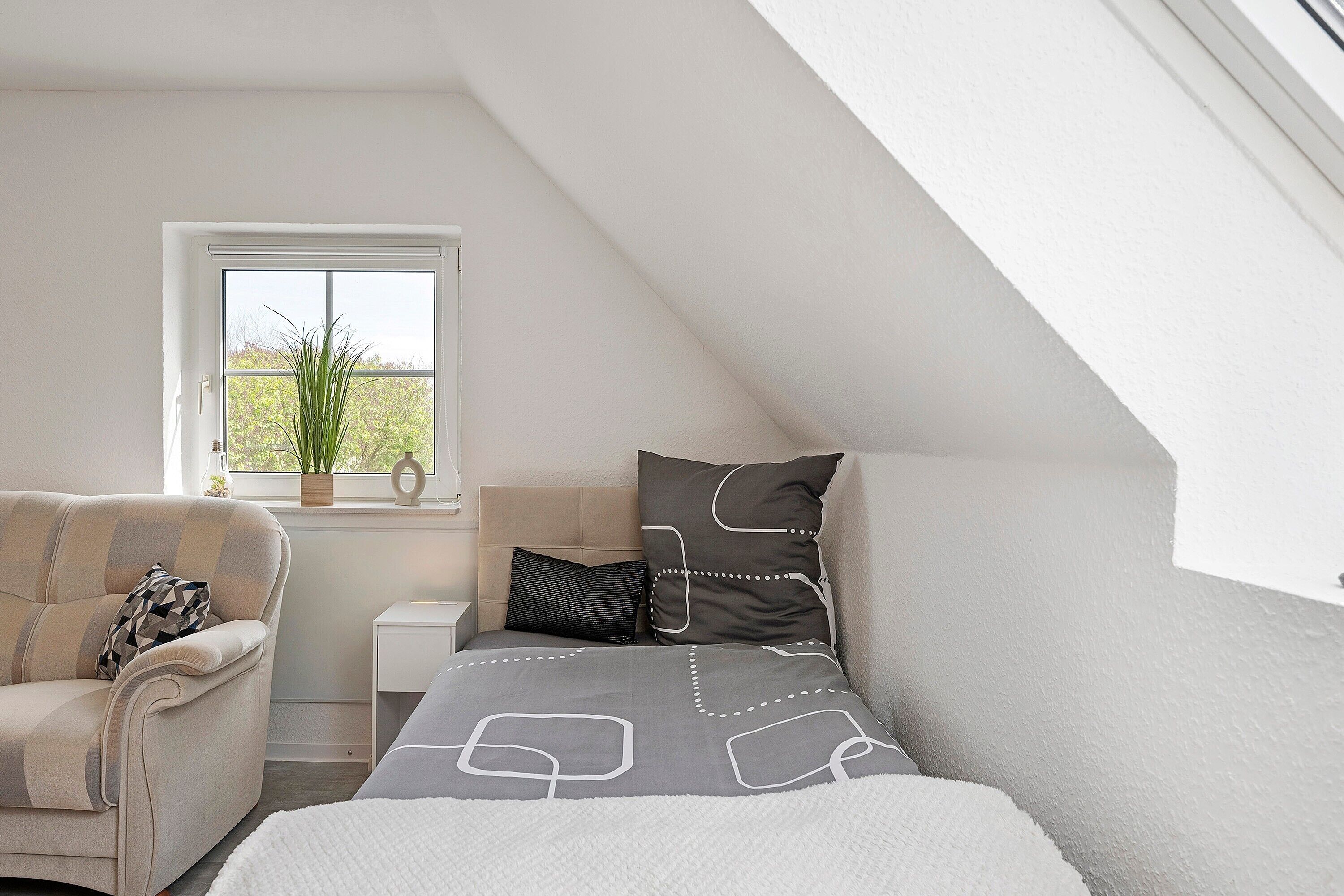 A bedroom with a bed, a chair, a nightstand, a window, and a plant.