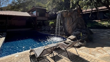 Una piscina con caída de agua, sillas de descanso y un edificio con tejado de baldosas.