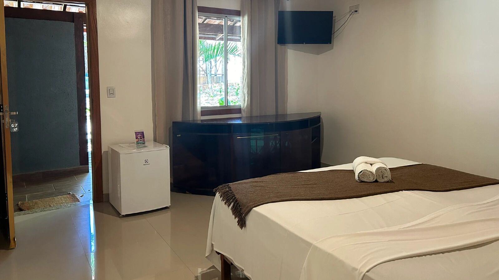 A hotel room with a bed, a small white refrigerator, a dark-colored cabinet, a television mounted on the wall, and a window with curtains.