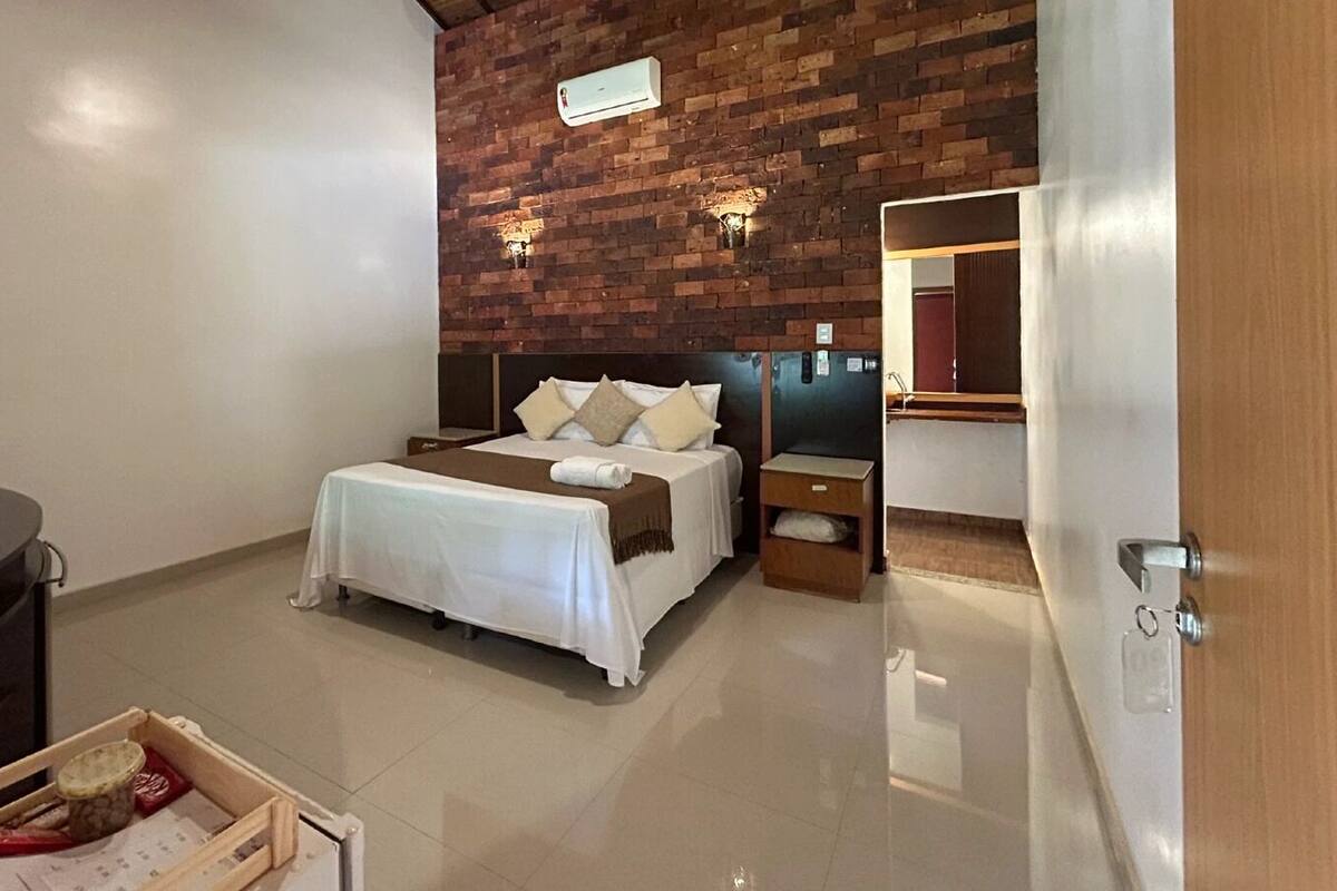 A hotel room with a brick accent wall, a bed with white bedding, a nightstand, a dresser, and a mirror.