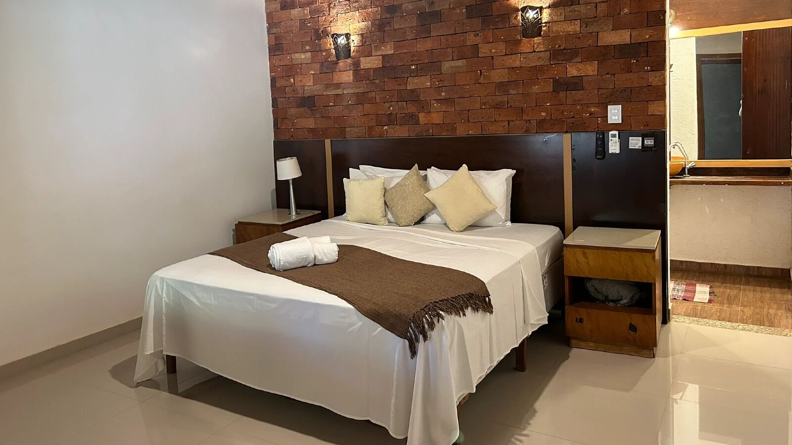 A hotel room with a brick wall, a bed with white linens, a nightstand, a wooden shelf, and a mirror.