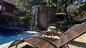 Piscina com cachoeira, espreguiçadeiras e um edifício com telhado de azulejos.