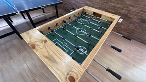 A wooden foosball table with a soccer field layout, a ping pong table, and a brick wall in the background.