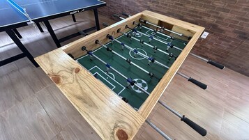 Uma mesa de futebol de madeira com design de campo de futebol, uma mesa de pingue-pongue e uma parede de tijolos ao fundo.