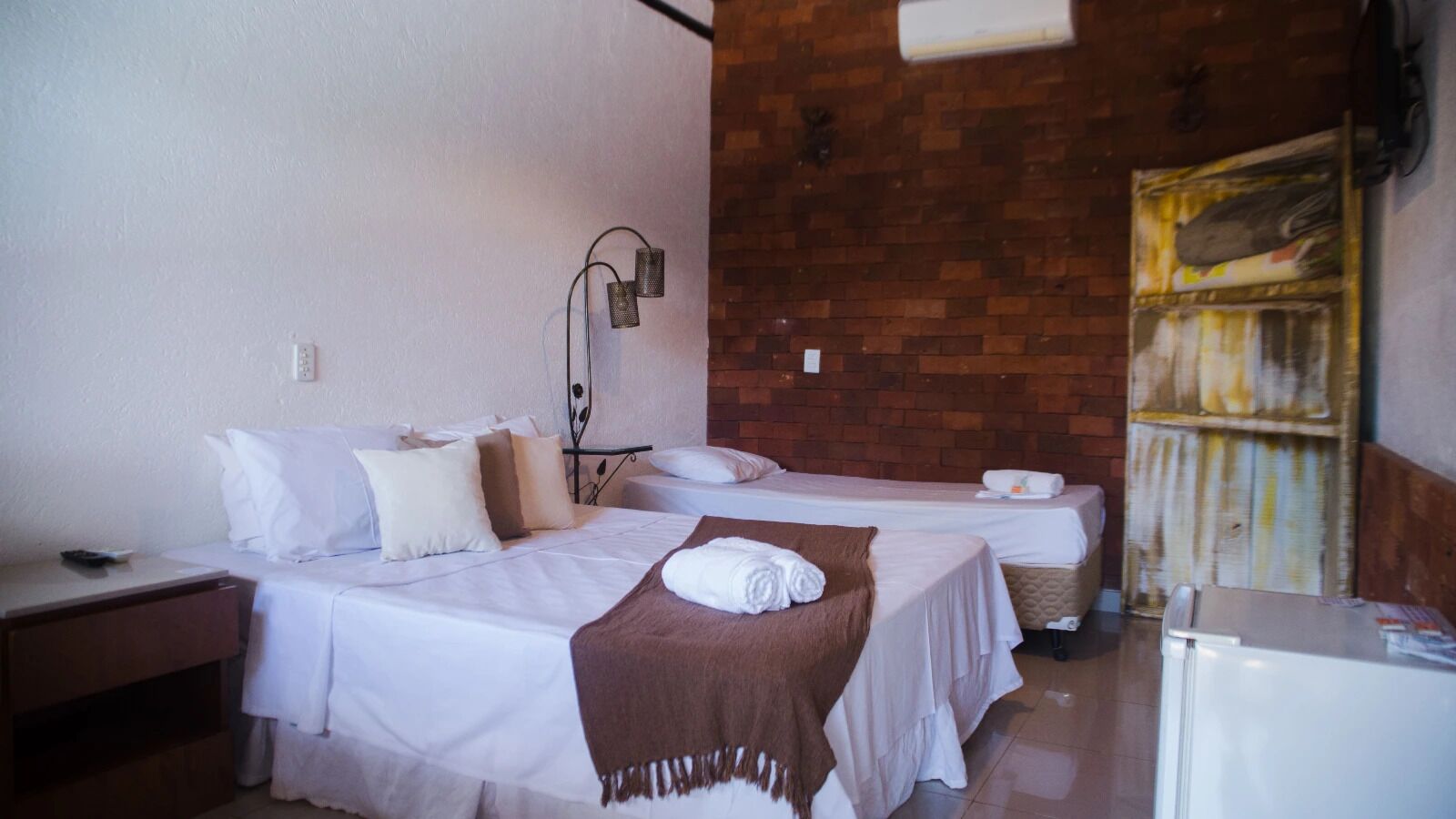 Une chambre d’hôtel avec deux lits, une petite table, une table de chevet, une lampe fixée au mur et un mur en briques.