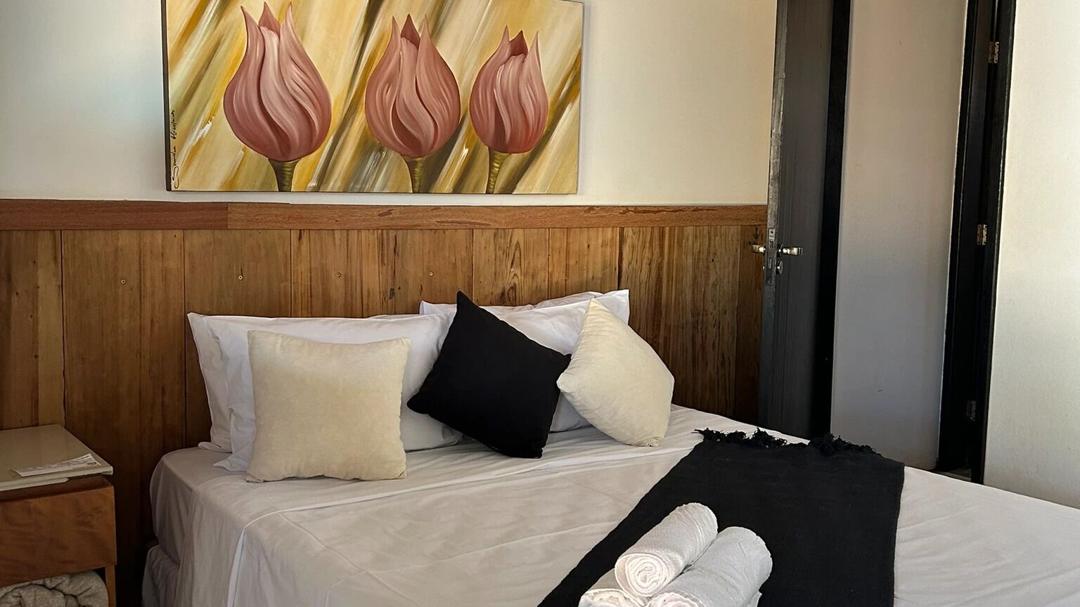 Habitación de hotel con cabecera de madera, cama con ropa blanca y un cuadro de flores en la pared.