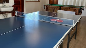 A ping pong table with two red paddles, a wooden pool table, and a sink in a room with wooden flooring.