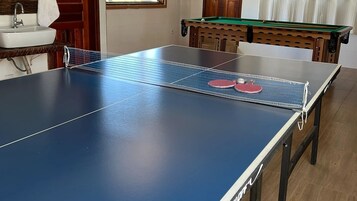 A ping pong table with two red paddles, a wooden pool table, and a sink in a room with wooden flooring.